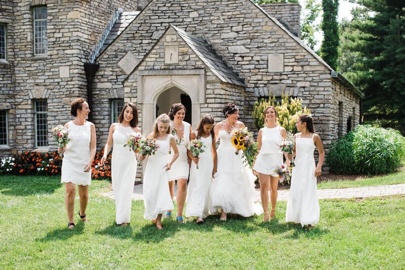Bride walking with bridesmaids and flower girls in white dresses holding bouquets outside a stone chapel, coordinated by Michaelis Events – Top Event Planners in Louisville KY & Best Wedding Caterers.