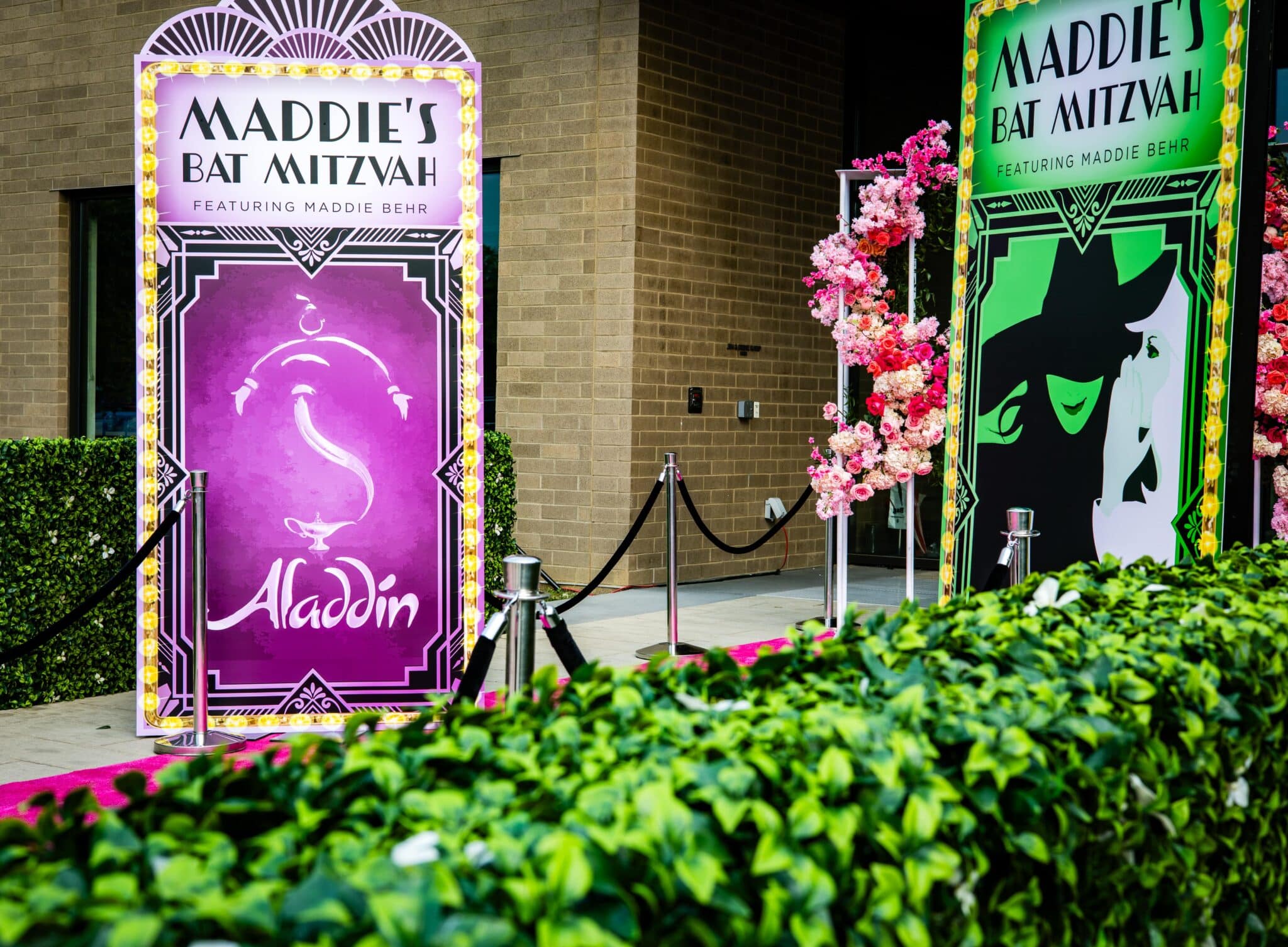 Broadway-themed entrance signs for Maddie's Louisville Bat Mitzvah, featuring custom “Aladdin” and “Wicked” posters with floral installations and velvet ropes. Designed by Michaelis Events. Captured by TIMB Photography.